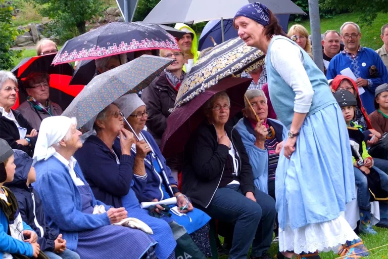 Frau in hellblauer traditioneller Tracht beugt sich zu einem, im Freien sitzenden Publikum herunter. Das Publikum besteht aus älteren Menschen, von denen einige ebenso Trachten tragen und Regenschirme über sich halten.