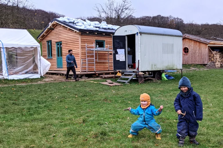 Kinder in Schneeanzügen spielen auf einer Wiese vor einem Holzhaus und einem Bauwagen